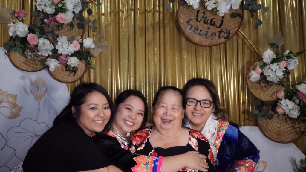 Three Hmong sisters hugging their elderly mother, wearing ethnic Hmong clothes. A sign "Vang Soiety" is shown in the back.