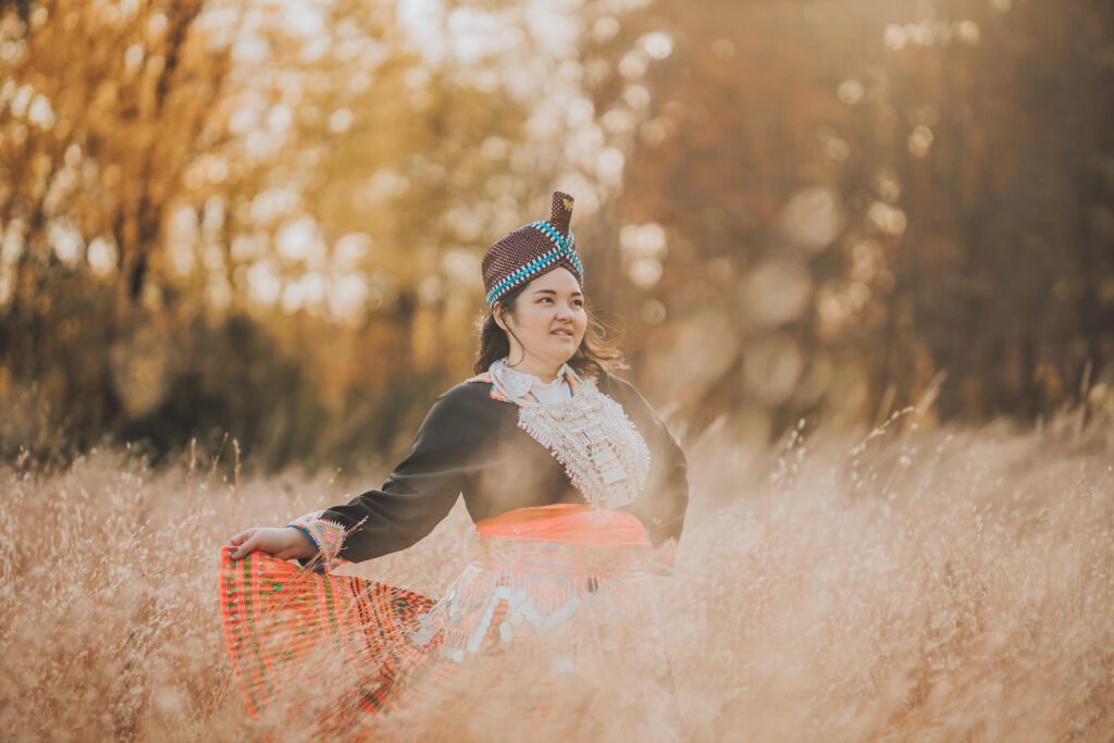 Mi Ong in traditional hmong clothes, with a purple glitter turban hat, black shirt with a heavy silver necklace, pink silk belt and a belt with beaded coins, and a colorful patterned skirt.